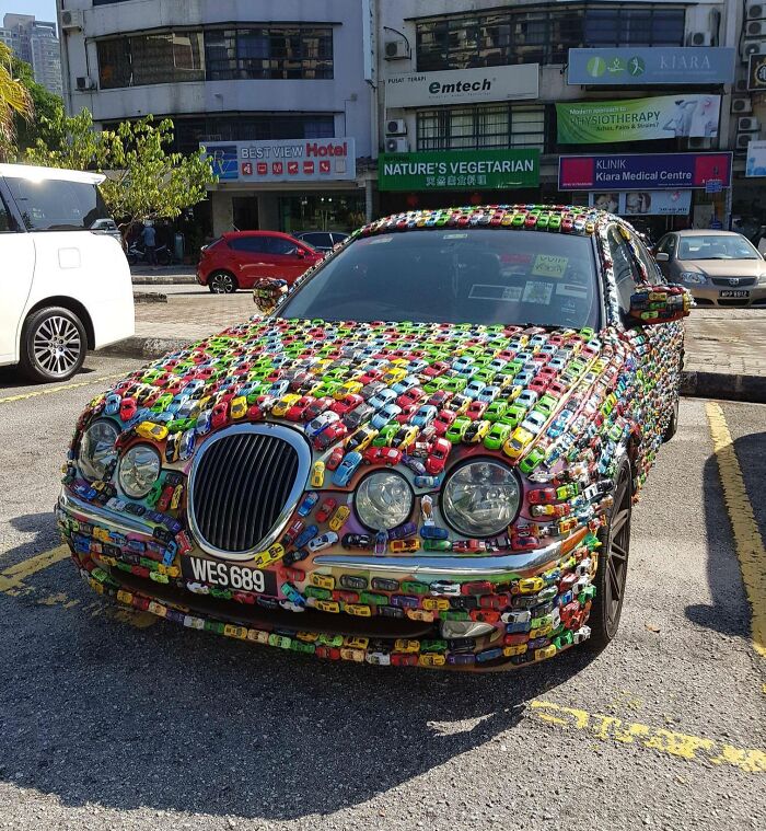 A car covered entirely in colorful toy cars, parked in an urban setting, illustrating a humorous car-fails moment.