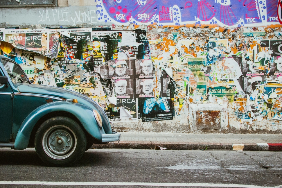 A vintage blue car is parked on a street in Uruguay, where a wall covered in weathered posters and graffiti. The pavement shows signs of wear, adding to the urban, atmosphere.