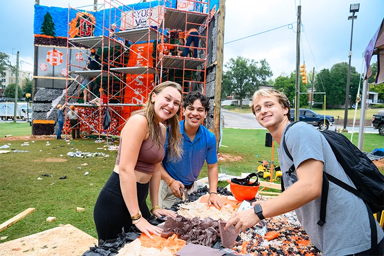 Three greeklife students work together on a part of a homecoming float display with other displays under construction in the background