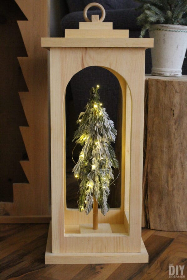 natural colored wooden lantern with faux tree sitting inside. 