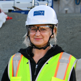 Portrait of a person with shoulder-length blonde hair, wearing a hardhat, safety goggles, and safety vest, smiling.