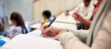 A closeup of a person's hand filling in an exam in a lecture hall with blurred forms of other exam takers in the background.