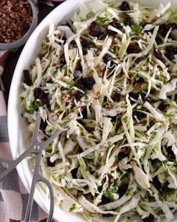 White bowl with salad tongs scooping portion of thin sliced green cabbage, dried cranberries, pecans and red onions.