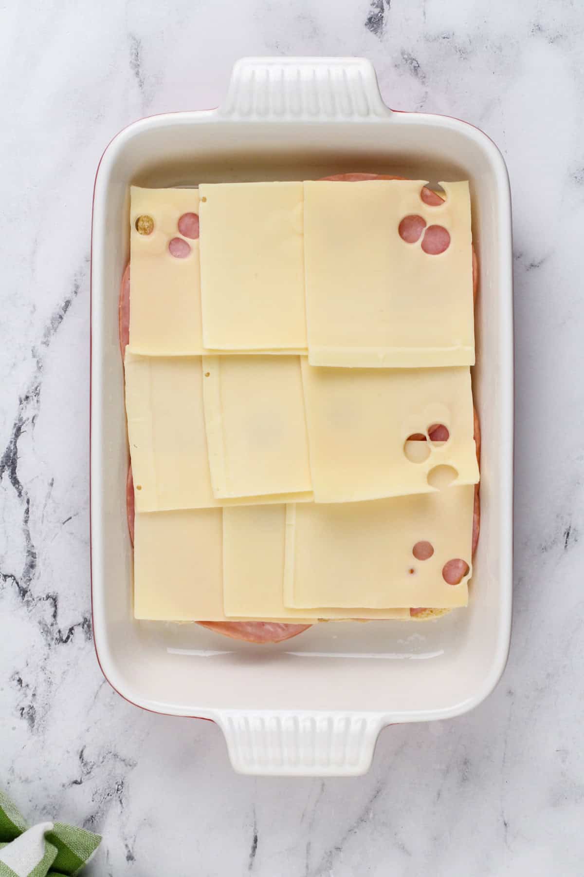 A layer of ham and swiss cheese layered on top of the bottom half of slider rolls in a ceramic baking dish.