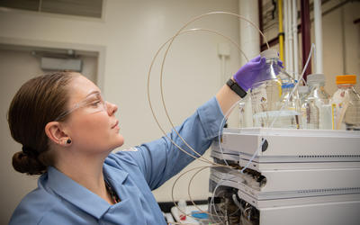 Alix Rodowa wears safety glasses and gloves in the lab as she reaches for a container of clear liquid on top of a piece of scientific equipment. 