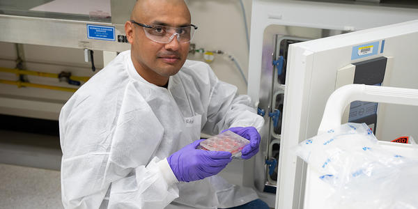 A researcher wearing safety gear kneels at the open door of a lab fridge, displaying a tray of samples. 