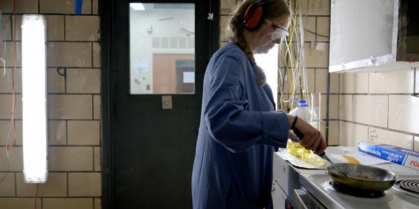 Side view of a woman wearing safety glasses, ear protection, and a blue lab coat. With one hand, she is holding a stainless steel frying pan over an electric stove. With her other hand, she is using a spatula to prod bacon in the pan. The room is smoky.