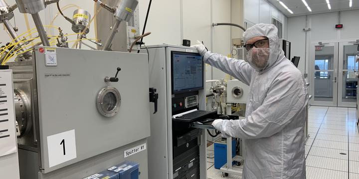 A person in a white coverall and safety glasses poses standing next to a rack of computers and other devices in a lab. 