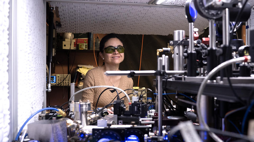 Ana Maria Rey wears dark safety glasses as she stands behind a table covered with optics equipment in the lab. 