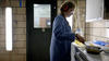 Side view of a woman wearing safety glasses, ear protection, and a blue lab coat. With one hand, she is holding a stainless steel frying pan over an electric stove. With her other hand, she is using a spatula to prod bacon in the pan. The room is smoky.