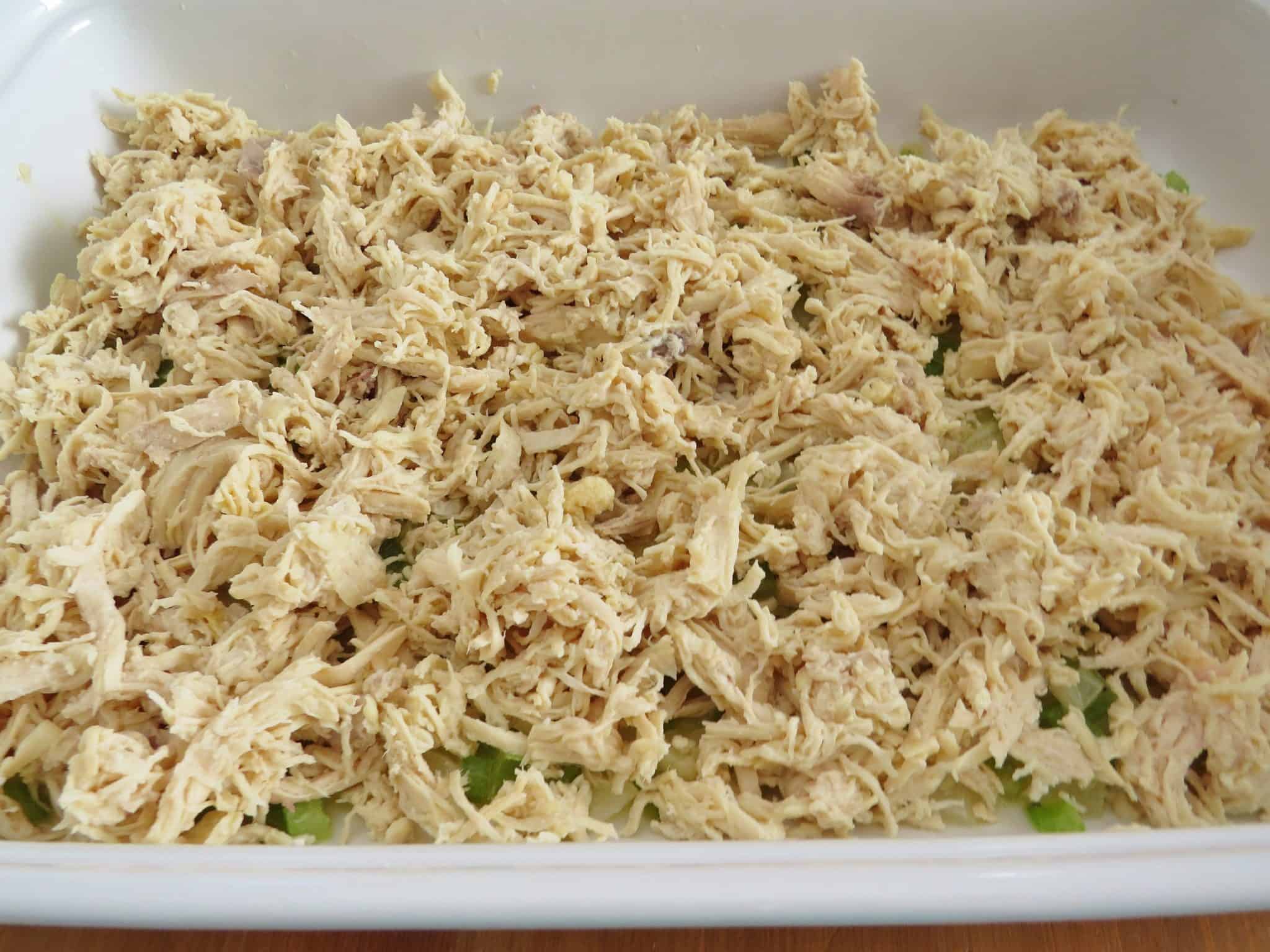 cooked, shredded chicken layered on top of malted butter, Clery and onion in a white baking dish.