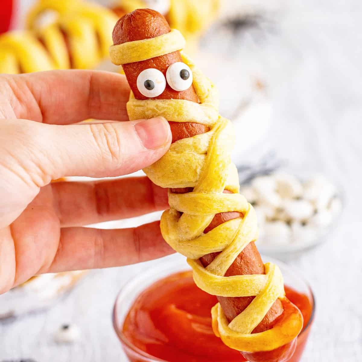 Close up of hand holding up one Halloween Mummy Dog dipped in ketchup.