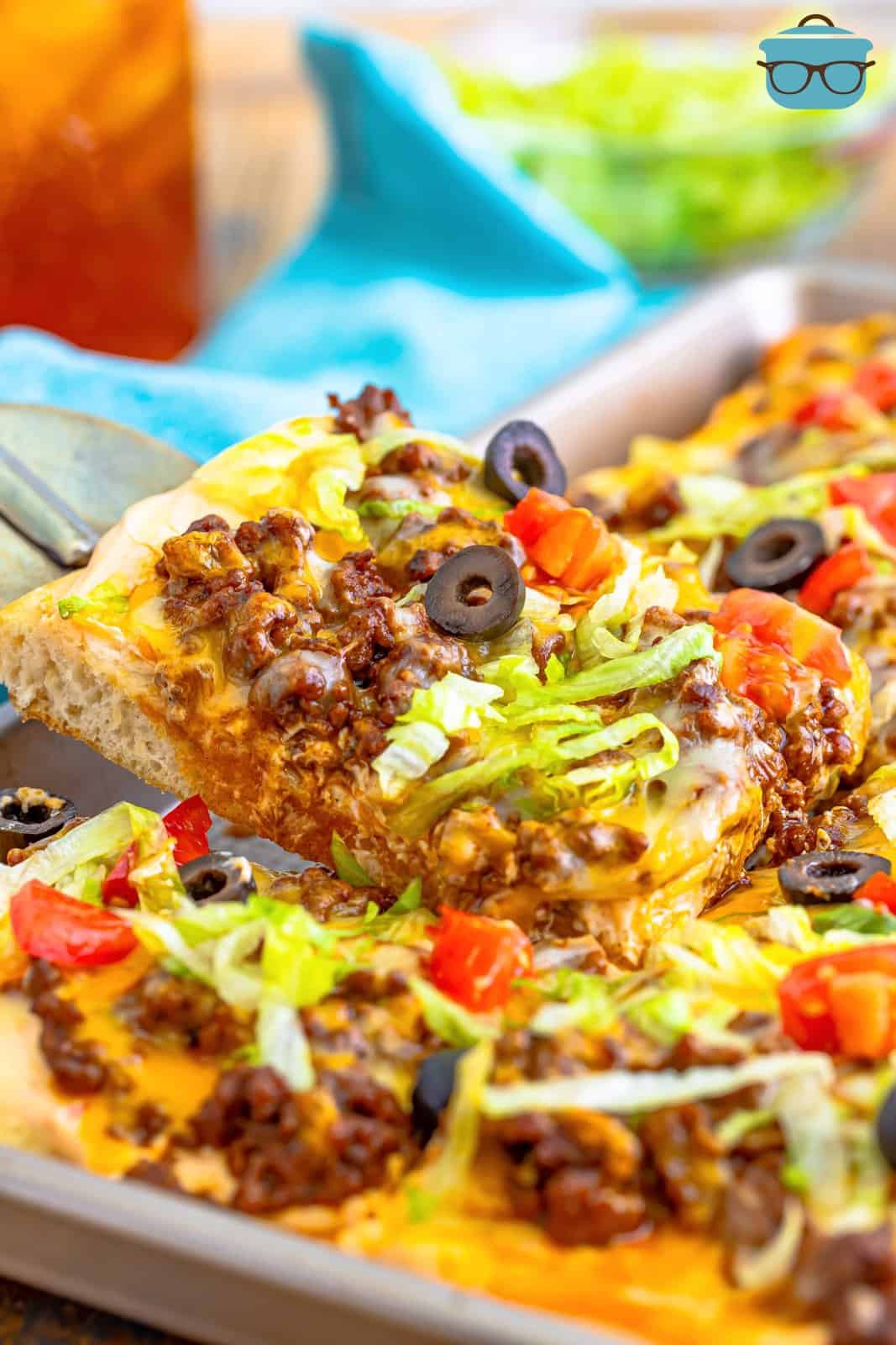 a spatula lifting up a square slice of Taco Pizza from a baking pan. 