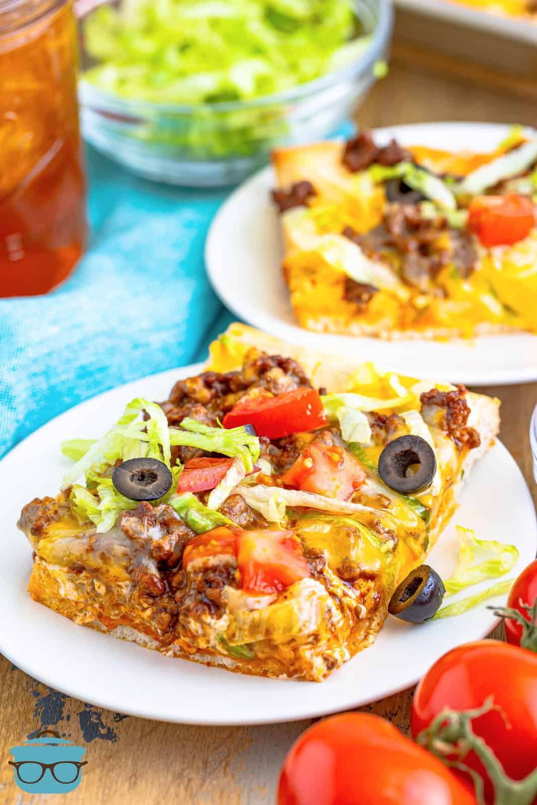 slice of taco pizza shown on a round white plate wit ha glass of iced tea in the background.