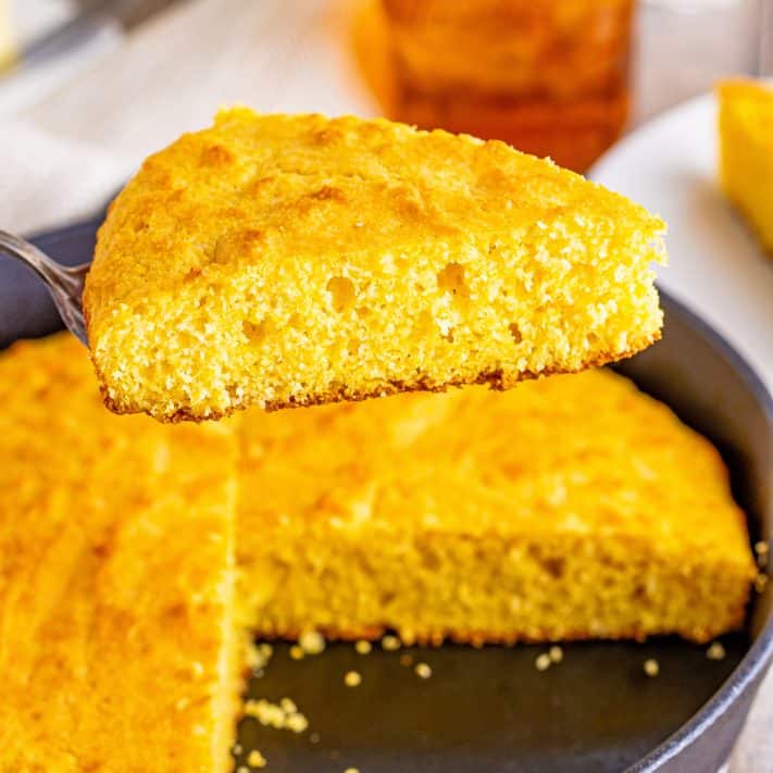 A slice of cornbread over a skillet filled with the rest of the loaf of cornbread.